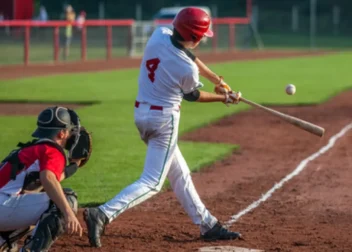 Baseball batter in white and red swings at a pitched ball near home plate while the catcher crouches behind.