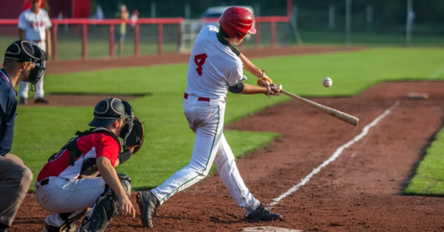 Baseball batter in white and red swings at a pitched ball near home plate while the catcher crouches behind.