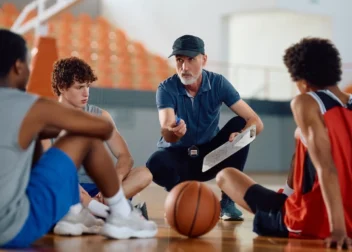 Basketball coach kneels on the court, clipboard in hand and whistle around his neck, teaching a small group of players.