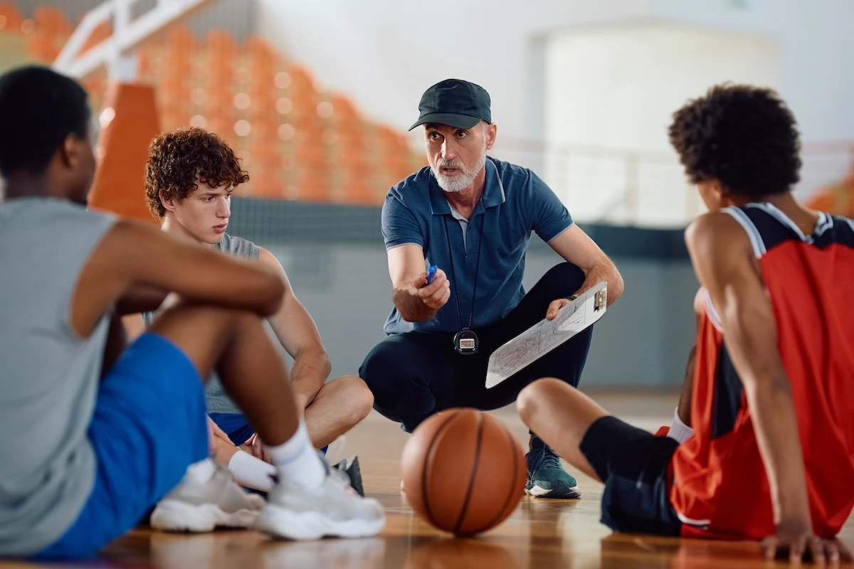 Basketball coach kneels on the court, clipboard in hand and whistle around his neck, teaching a small group of players.